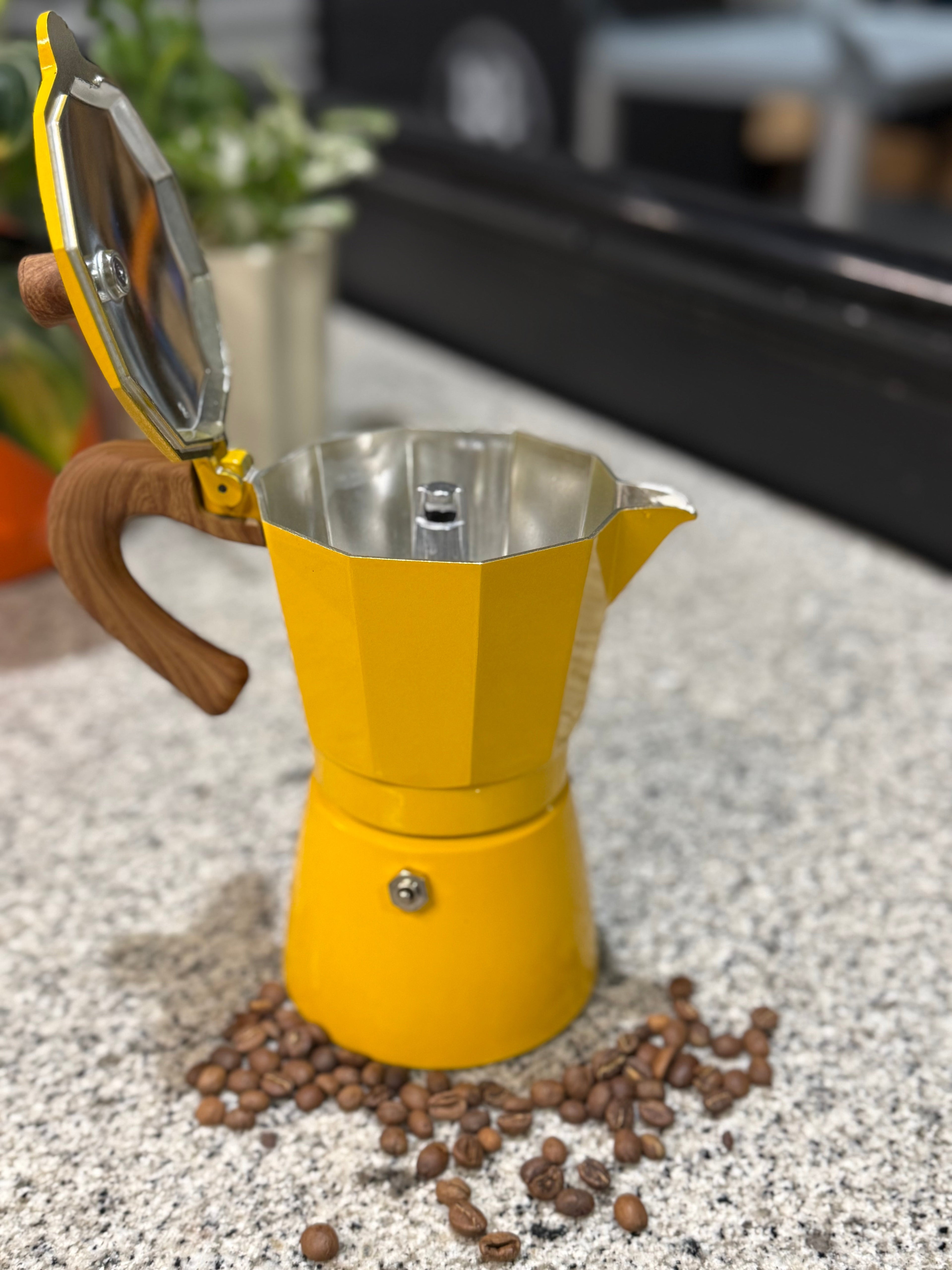 Yellow espresso maker with wooden handle on a speckled countertop with coffee beans around it.
