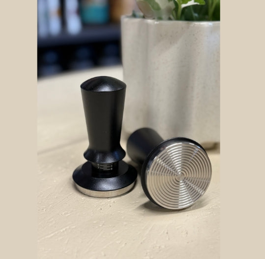 Two black coffee tampers on a light surface with a blurred plant in the background